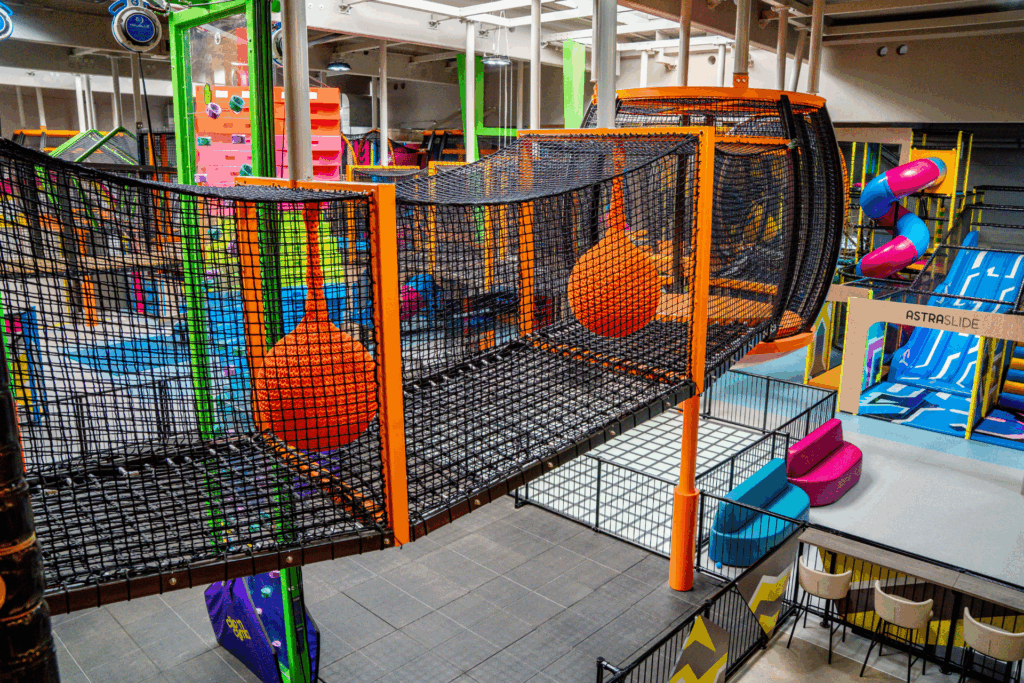 Ein drei Meter hohes Höhenlabyrinth in einer Indoor Halle. Die Umrandung ist orange und das Netz schwarz. In dem Labyrinth sind verschiedene Hindernisse. Im Hintergrund sind blau pinke Rutschen.