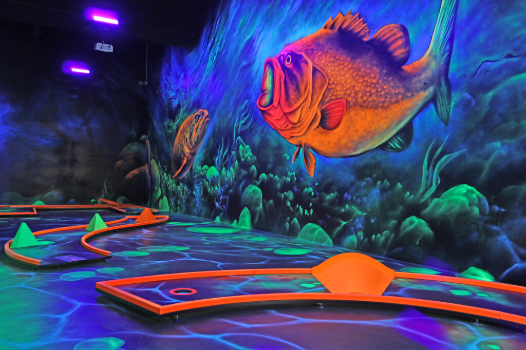 Bei der 3D Blacklight Golf Spielbahn handelt es sich um das Thema Unterwasserwelt. Dabei ist ein großer neonoranganer Fisch an der Wand abgebildet und danbenen eine Unterwasserwelt aufgezeichnet.