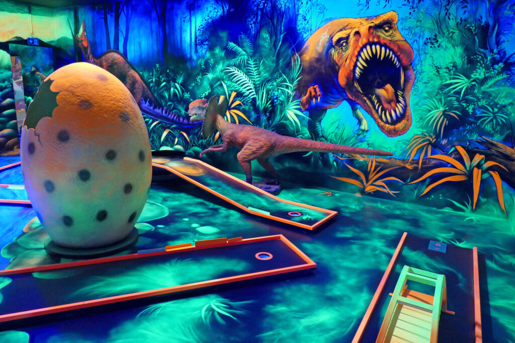 Bei der 3D Blacklight Golf Spielbahn handelt es sich um das Thema Dinosaurier. Es sind Dinosaurier Figuren im Vordergrund aufgestellt und die Wand ist mir Dinos bemalt.