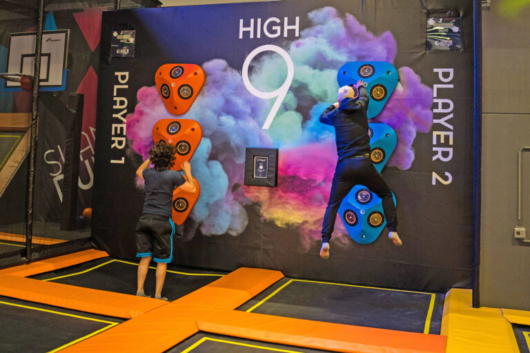 Zwei Männer springen auf zwei Trampolinen in die Luft und drücken Buzzer auf der Kletterwand, wichtig ist dass sie schnell reagieren um die Buzzer zu drücken.