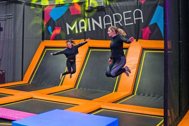 Zwei junge Frauen, welche schwarz gekleidet sind und Sportklamotten tragen springen auf einer großen Trampolinfläche in einer Trampolinhalle.