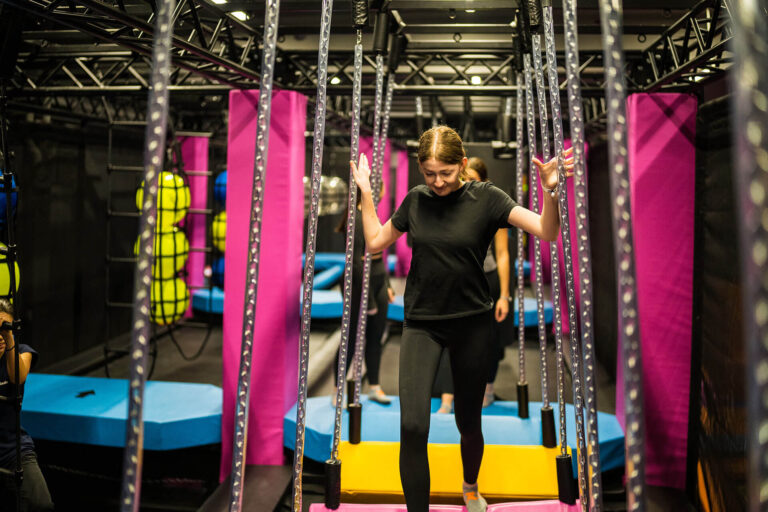 Ein braunhaariges Mädchen geht über wackelige farbige Balken, welche mit Seilen festgemacht sind. Sie befindet sich in einem Ninja Parcours.