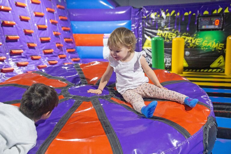 Zwei kleine Kleinkinder sitzen auf dem Wibble Wobble Bed in dem lila Inflate Park und erkunden diesen.