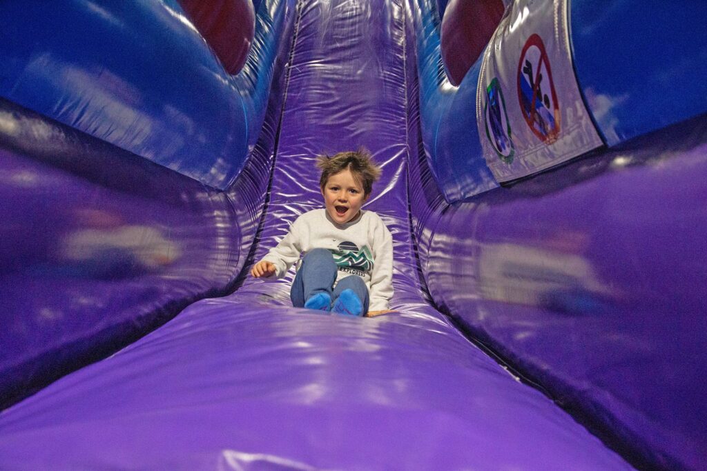 Ein kleiner Junge rutscht eine lila farbene Rutsche runter, welche Teil eines Inflate Parks ist. Seine braunen Haare sind leicht elektrisiert.