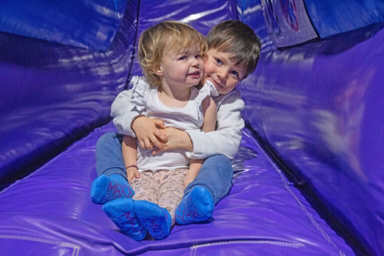 Ein kleiner Junge und ein kleines Mädchen umarmen sich und lächeln dabei. Sie sitzen auf einer Rutsche in einer lila farbenen Hüpfburg.