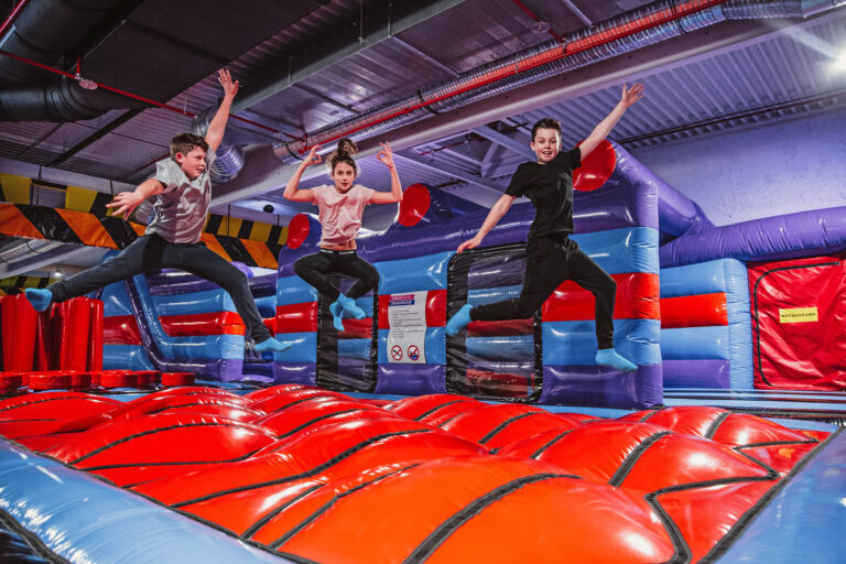 Drei Kinder, zwei Jungs und ein Mädchen, springen in die Luft auf einem riesigen roten Luftkisssen. Sie befinden sich in einem Inflate Park.