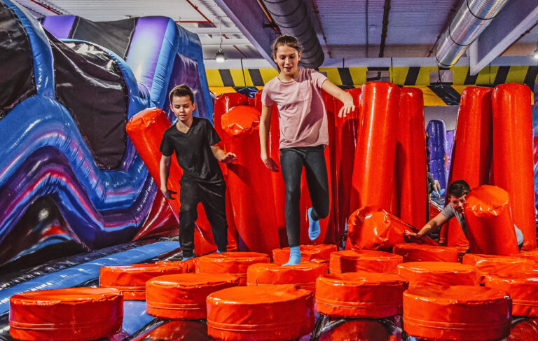 Kinder spielen im Inflate Park. Dabei spielen sie in der Agility Zone, wobei sie über rote weiche Hinternisse klettern.