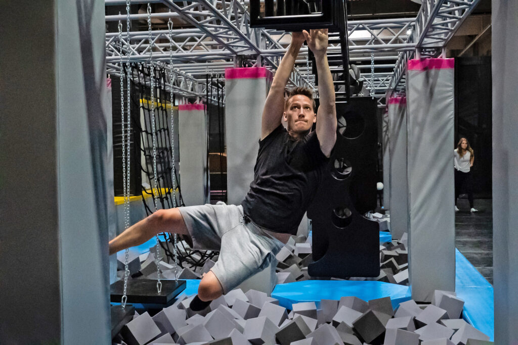 Ein erwachsener Mann mit schwarzem T-Shirt hangelt sich durch einen Ninja Parcours. Er schwebt in der Höhe und ist mitten im Parcours.