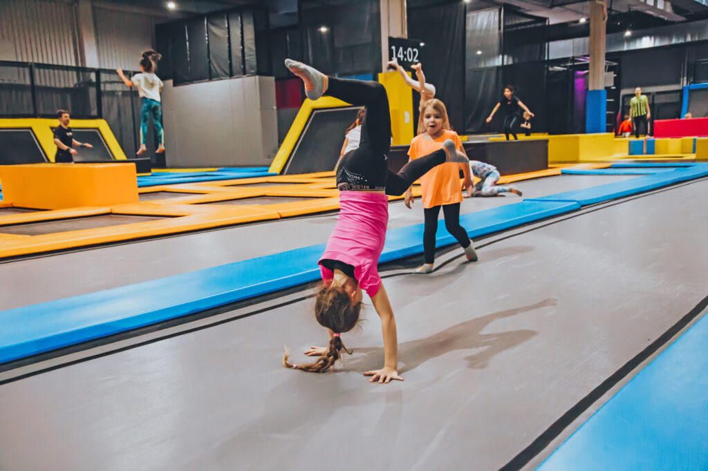 Ein kleines Mädchen mit einem pinken T-Shirt befindet sich auf einem länglichen Trampolin und macht einen Salto. Im Hintergrund sind weitere Kinder zu sehen.