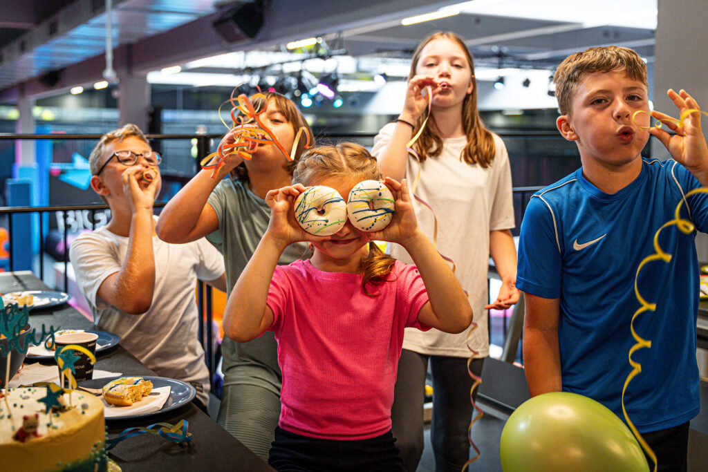 Ein Mädchen hält sich zwei Donuts vor ihre Augen, während vier Kinder im Hintergrund Luftschlangen aufpusten.