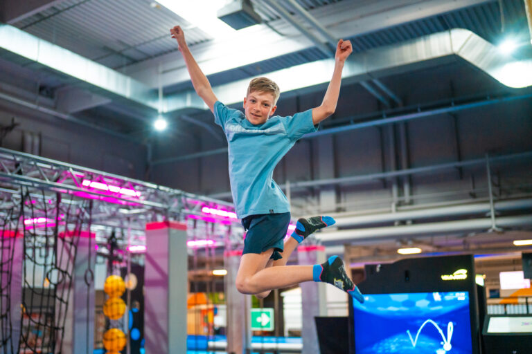Ein Junge in türkisenen T-Shirt und Hose springt Trampolin und reißt die Hände in die Höhe während er lächelt.