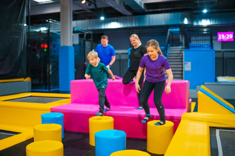 Eine junge Familie mit zwei Eltern und zwei Kindern sind auf der Trampolinfläche. Die Kinder springen über einen Parcour, während der Vater und die Mutter hinter den Kindern gehen.