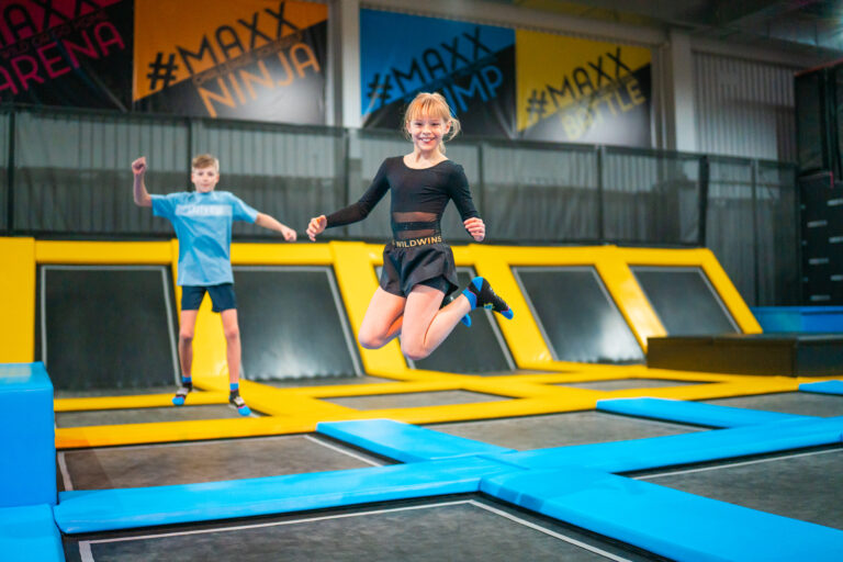 Ein kleine blondes Mädchen springt Trampolin, zieht dabei ihre Füße an und streckt die Arme leicht in die Höhe. Sie lächelt und ist glücklich. Im Hintergrund springt ebenso ein kleiner Junge Trampolin