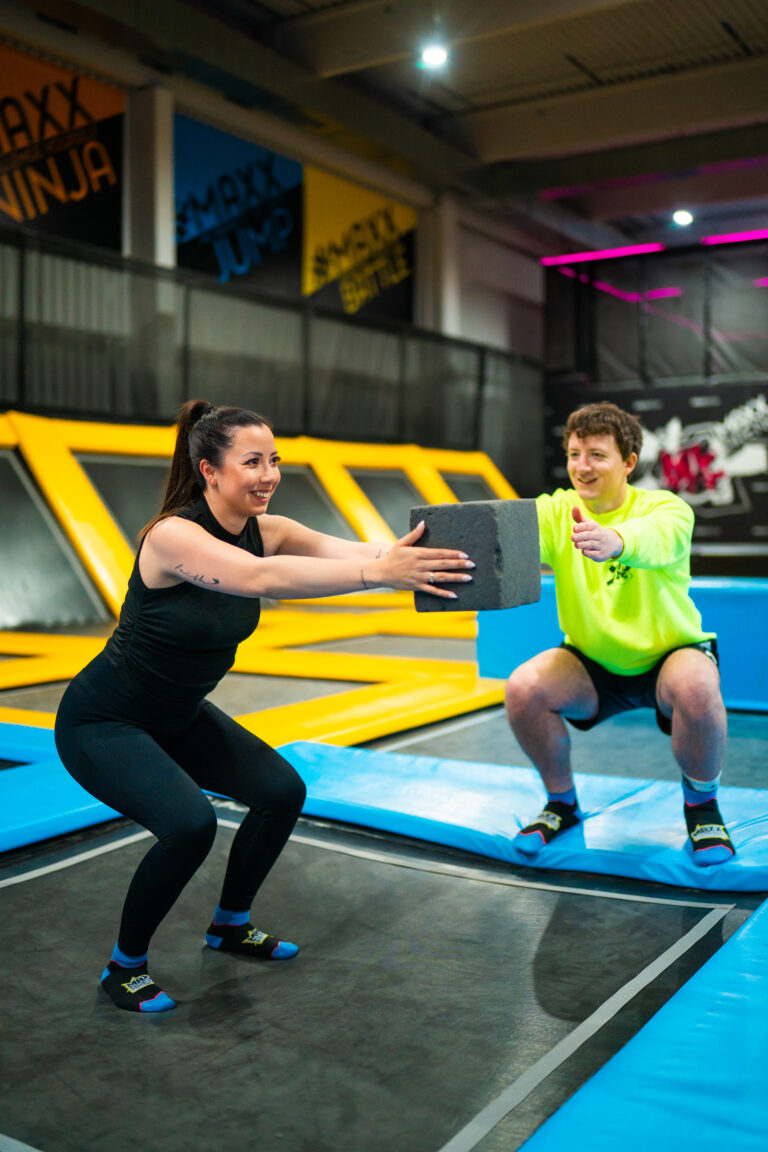 Eine Frau führt eine Fitnessübung in einer Indoor Trampolinhalle namens Maxx Arena durch. Die Frau hält einen Schaumstoffblock in ihren ausgestreckten Armen, während sie und ein Trainer in der Hocke stehen.